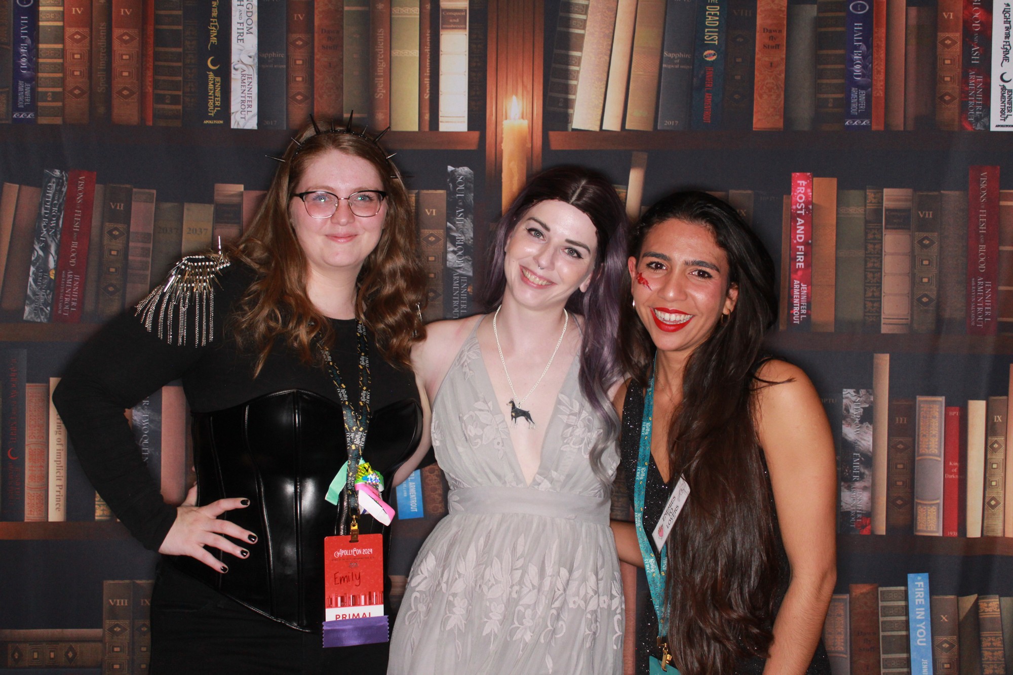 Three friends posing against the bookshelf backdrop designed for ApollyCon